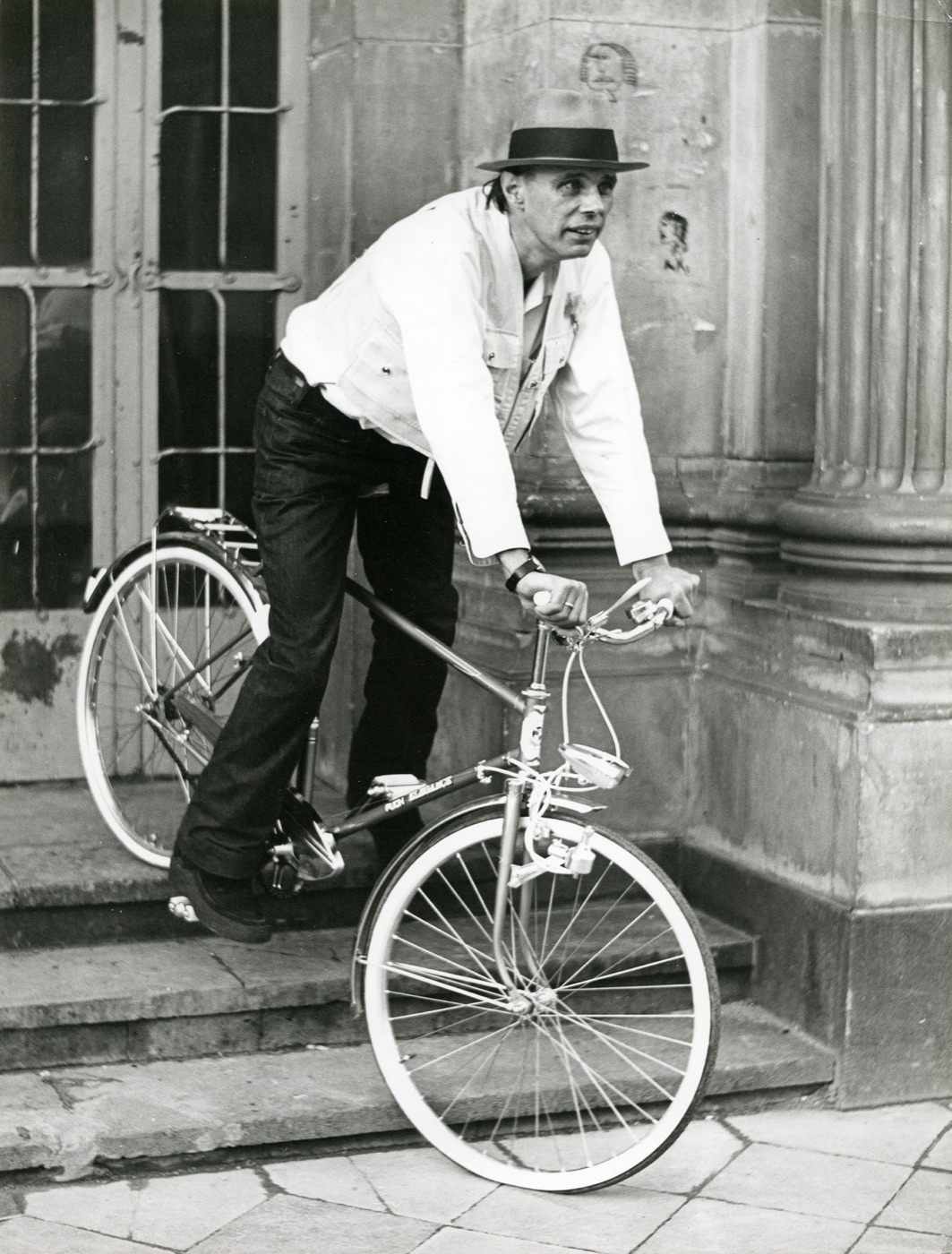 Joseph Beuys en chapeau, poussant un vélo dans un escalier de pierre