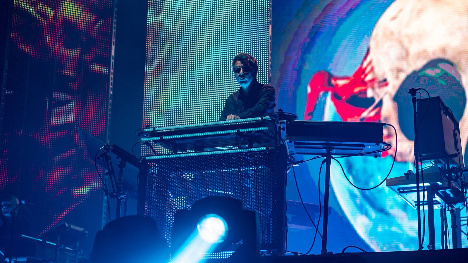 Jarre in sunglasses at a rig of keyboards, before a huge screen of colored images