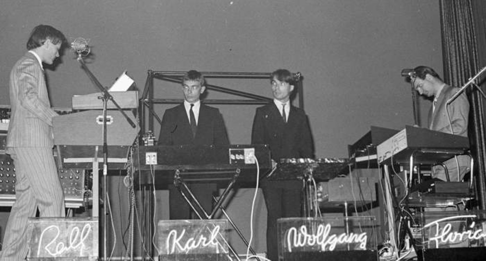 Quatre hommes en costume aux claviers sur scène, derrière des néons affichant les noms Ralf, Karl, Wolfgang et Florian