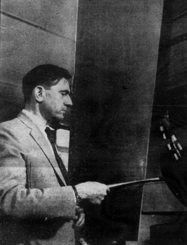 Pierre Schaeffer in a jacket and tie, holding a stick against a tall metal sheet