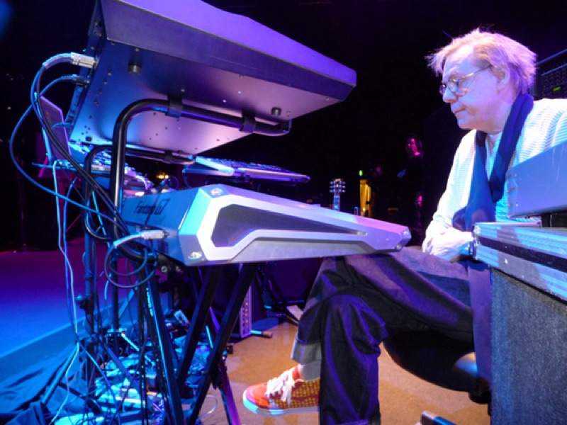 Klaus Schulze, white-haired, seated at a stack of keyboards under blue light