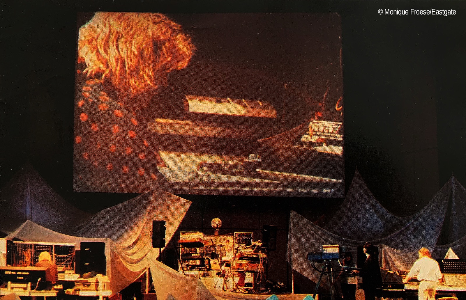 A concert stage with keyboards beneath a large screen showing a close-up of a musician