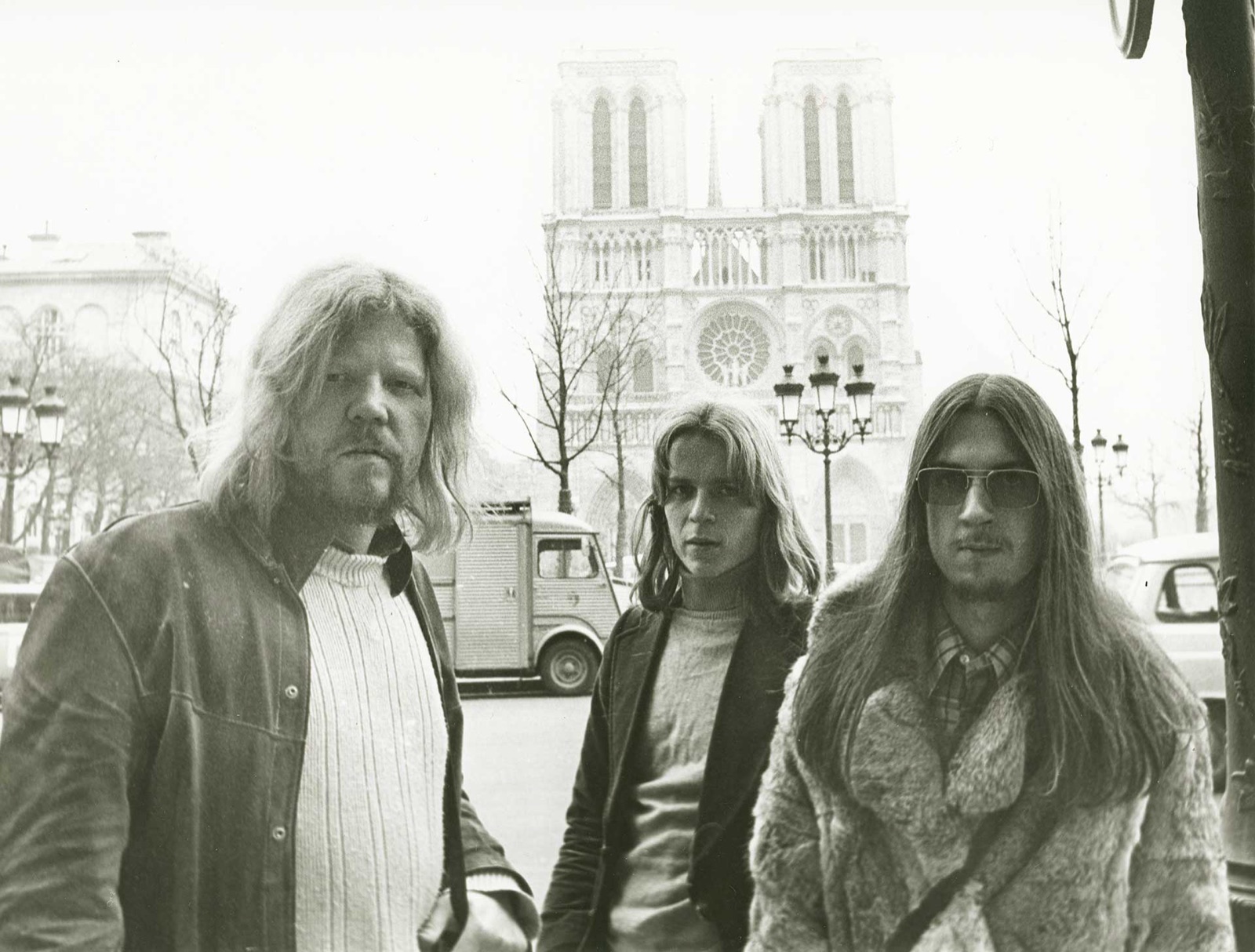Three long-haired young men in a Paris street, with the towers of Notre-Dame behind them