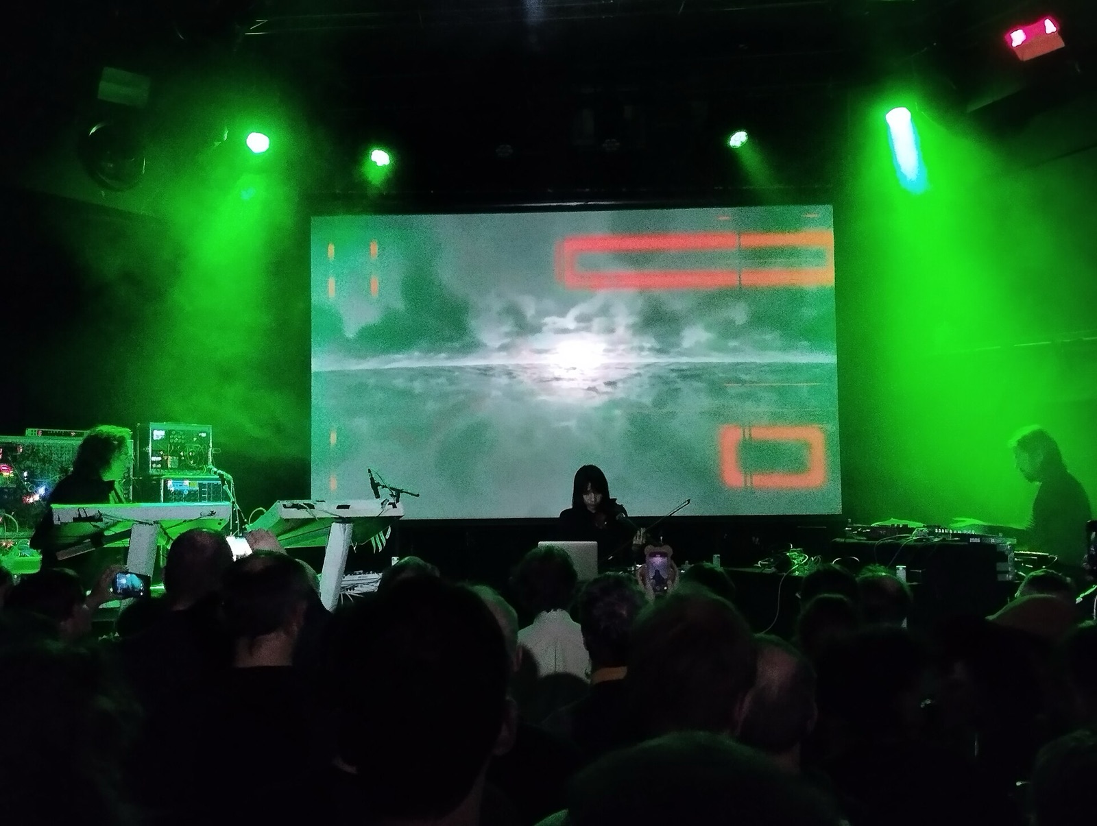 Eine dunkle Clubbühne mit Musikern an Keyboards unter einem großen projizierten Bild